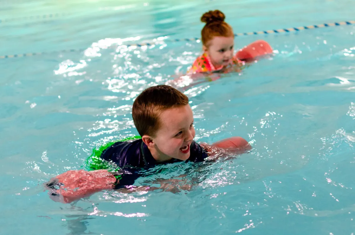 Two young Superfish swimmers kicking across the pool with foam noodles
